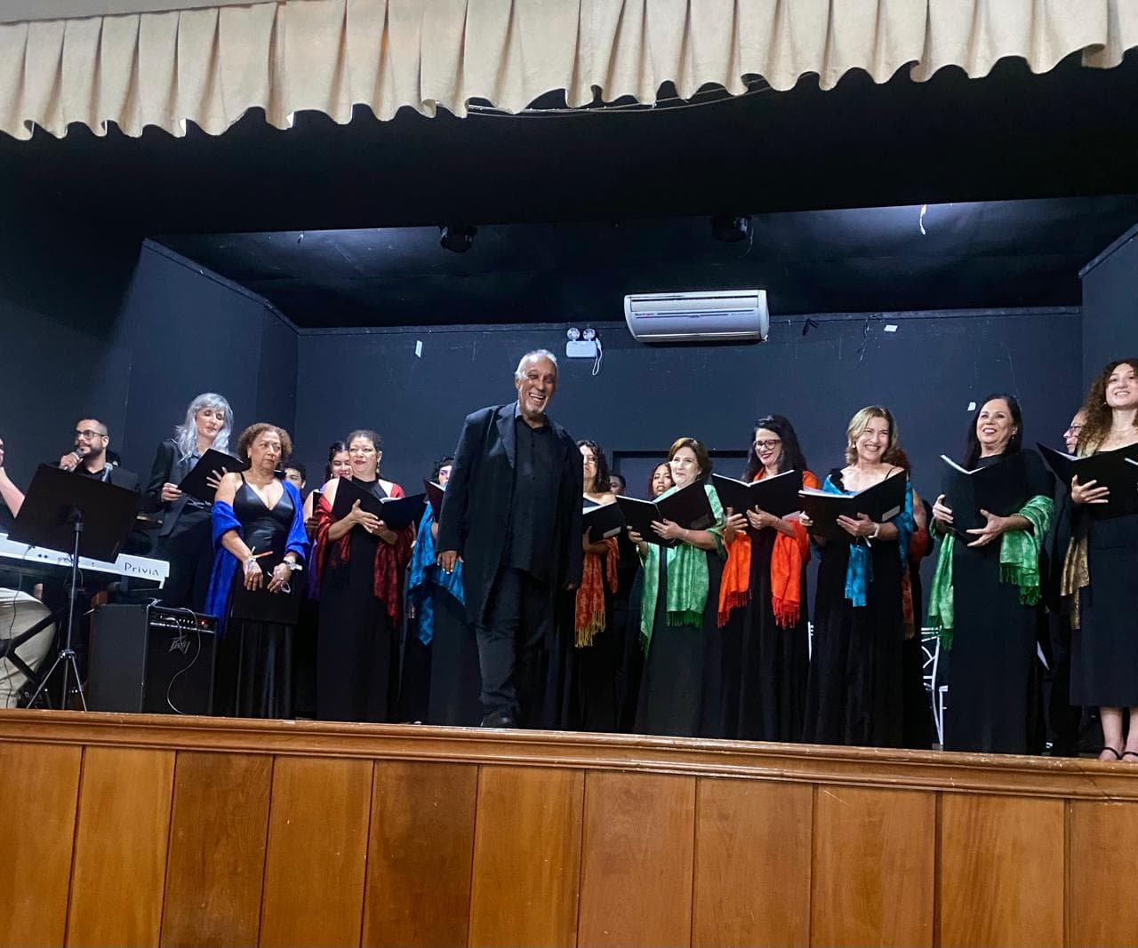 Maestro e coralistas sorrindo no palco ao fim de uma apresentação